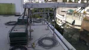 cavalete com contrapreso Estrutura metálica de suporte com contrapesos empilhados, utilizada para sustentar andaimes suspensos em obras. Fios e cabos estão espalhados no chão ao redor da estrutura, e prédios residenciais aparecem ao fundo.