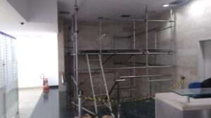 Imagem de um andaime montado no interior de um prédio, próximo a uma escada, para realização de serviços em altura. A estrutura metálica conta com plataformas de trabalho e escada de acesso, garantindo segurança aos trabalhadores. A área ao redor está sinalizada com fita de segurança para evitar circulação indevida. O ambiente possui paredes revestidas e balcão de recepção.