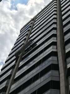 Imagem de um prédio alto com fachada envidraçada, onde um andaime suspenso (balancim) está instalado e pendurado por cabos de aço. O andaime é utilizado para trabalhos em altura, como manutenção ou limpeza da fachada. O céu parcialmente nublado aparece ao fundo faça seu orçamento
