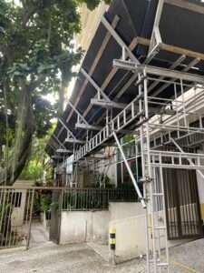 Imagem de um andaime aparalixo instalado em frente a um edifício residencial, garantindo proteção para pedestres contra queda de detritos durante a obra. A estrutura metálica conta com reforço e cobertura de madeira e lona. O equipamento é fornecido pela Nesfera Andaimes, especialista em soluções para trabalhos em altura.