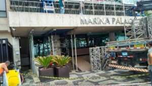 Imagem de uma obra em andamento na entrada do edifício Marina Palace, onde escoras metálicas e andaimes da **Nesfera Andaimes** estão instalados para sustentação temporária da estrutura. Trabalhadores realizam a montagem e ajuste dos equipamentos, enquanto um caminhão descarrega mais peças metálicas. A área ao redor está sinalizada com fitas de segurança, garantindo a proteção de pedestres.
