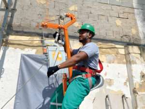 Trabalhador utilizando cadeira suspensa para realizar manutenção em uma fachada, equipado com capacete verde, luvas e cinto de segurança. A cadeirinha está presa a um sistema de cabos e roldanas, garantindo segurança durante a atividade em altura.