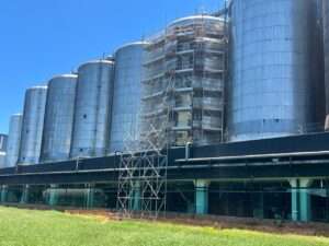 Grande estrutura metálica com vários silos cilíndricos de armazenamento. Em um deles, há um andaime multidirecional montado, cobrindo boa parte da superfície. O céu azul aparece ao fundo. No canto inferior esquerdo, um texto em destaque diz 'Andaime Multidirecional'.